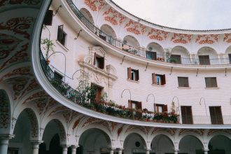 Pátio histórico em Sevilha, com arcadas ornamentadas, colunas e balcões ao redor de um espaço circular.