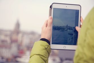 Mãos seguram um tablet diante de uma paisagem urbana, representando o uso da tecnologia na exploração turística.
