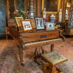 Piano antigo de madeira ornamentada em uma sala clássica, cercado por móveis e elementos decorativos.