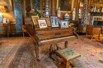 Piano antigo de madeira ornamentada em uma sala clássica, cercado por móveis e elementos decorativos.