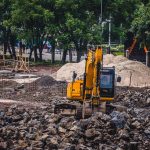 Escavadeira amarela trabalha em um canteiro de obras urbano cercado por árvores e pedras.