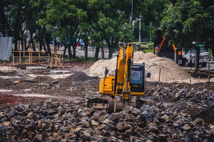 Escavadeira amarela trabalha em um canteiro de obras urbano cercado por árvores e pedras.