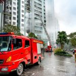 Bombeiros combatem a fumaça de um incêndio em um edifício residencial, com caminhões de emergência na rua.