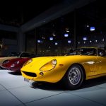 Carros esportivos clássicos alinhados em exibição dentro de um salão automotivo, com um modelo amarelo em destaque.