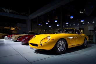 Carros esportivos clássicos alinhados em exibição dentro de um salão automotivo, com um modelo amarelo em destaque.