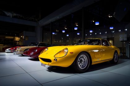 Carros esportivos clássicos alinhados em exibição dentro de um salão automotivo, com um modelo amarelo em destaque.